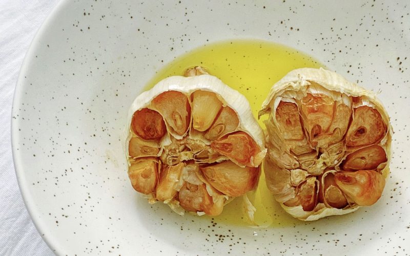 Roasted Garlic Recipe: a Step by Step to Roast Garlic in the Oven featured by top US food blog, Food Diary of a City Girl: image of a while bowl with black specks with two roasted garlic heads drizzled in olive oil.